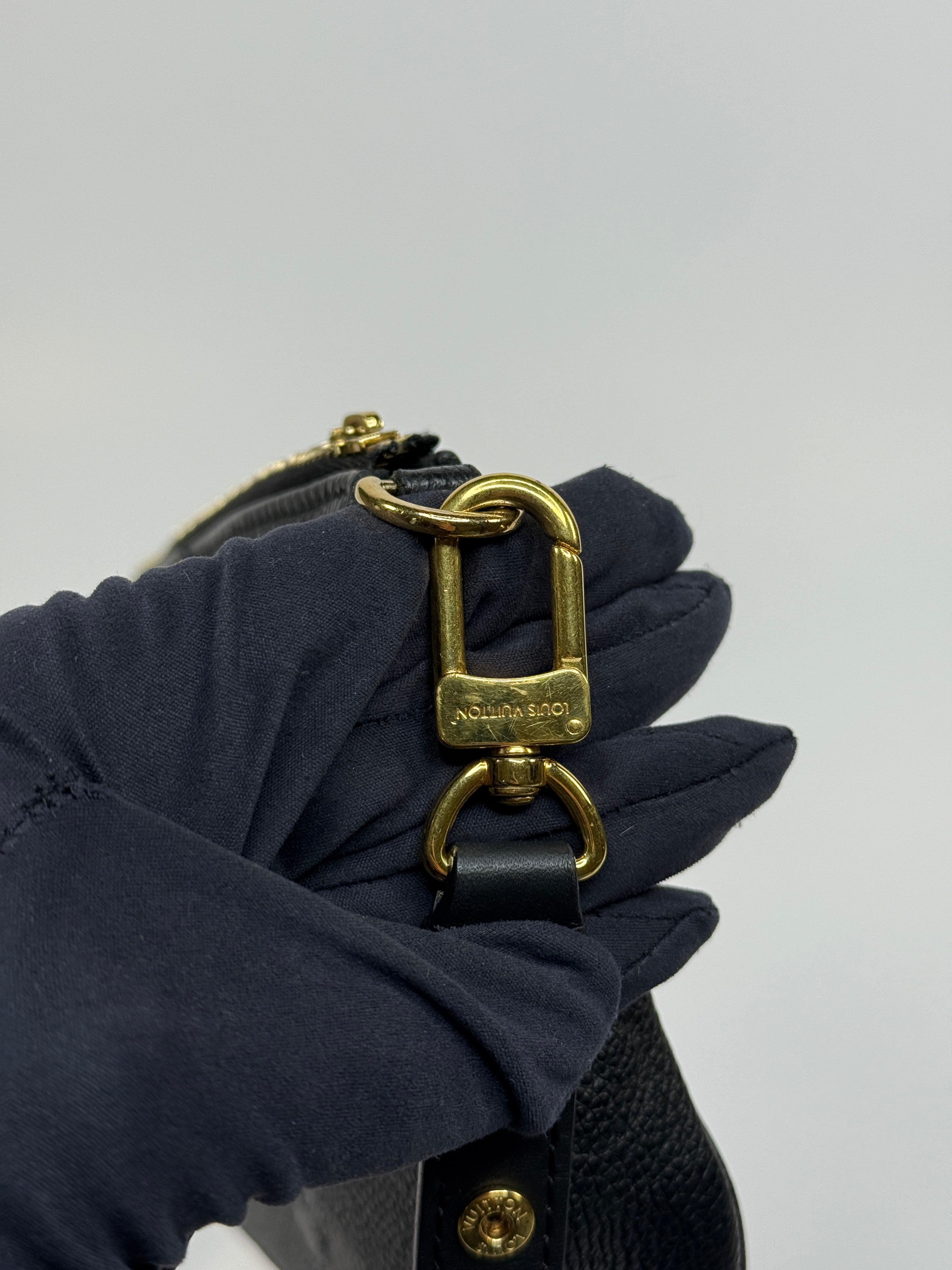 Close-up of a black sandal with gold buckle being worn by a gloved hand on a light gray background